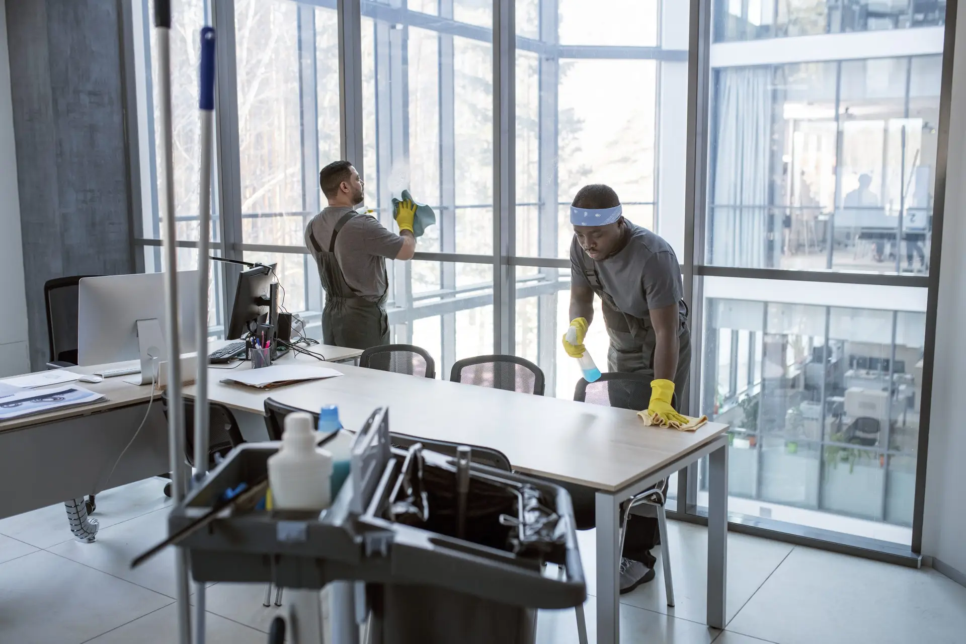 Zwei Mitarbeiter einer Reinigungsfirma reinigen ein modernes Büro, wischen Fenster und Schreibtische mit professionellen Reinigungsmitteln. Unterhaltsreinigung, Büroreinigung bei Erdan Glas- und Gebäudereinigung.