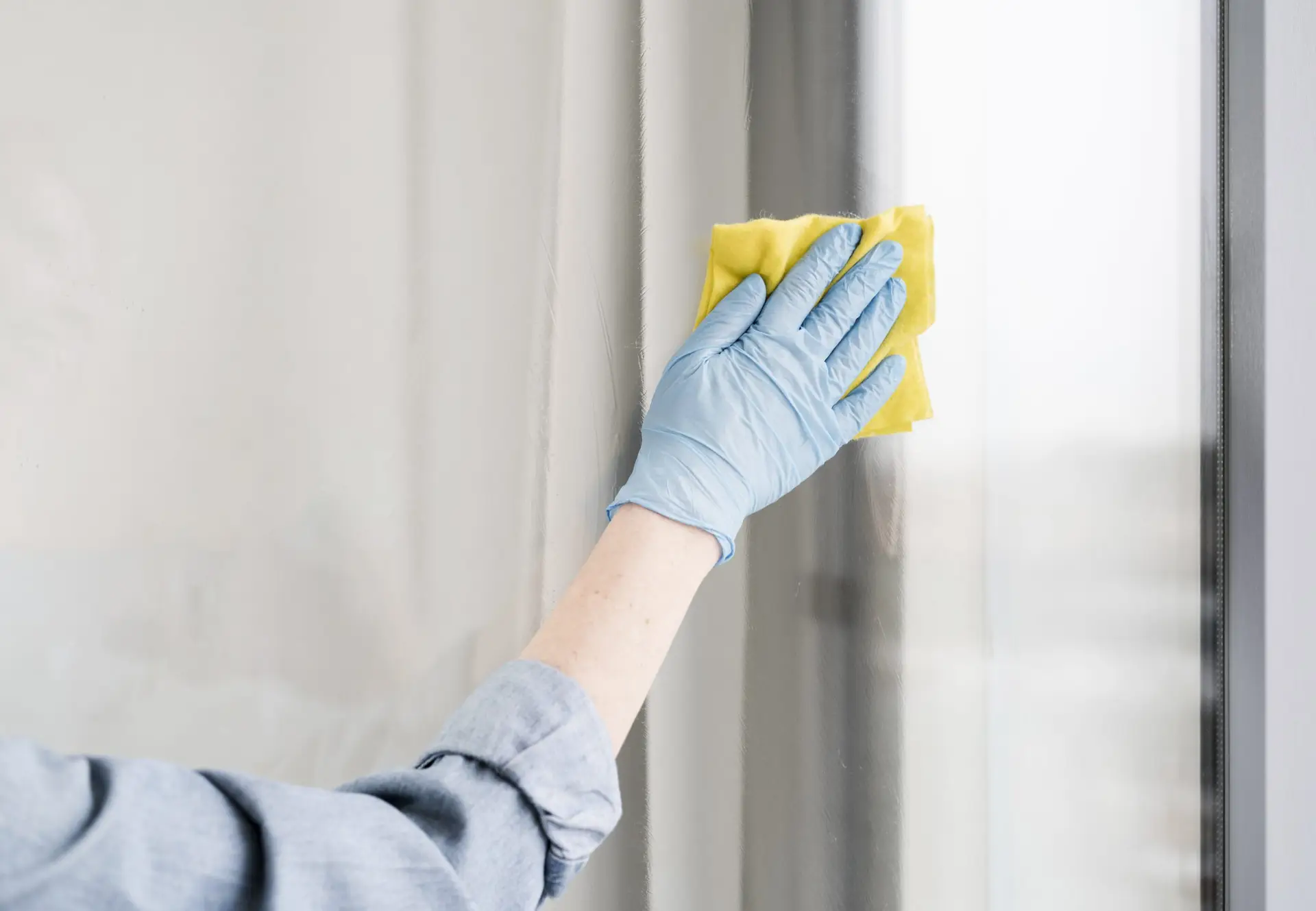 Eine Person mit blauer Einweg-Gummihandschuhen reinigt ein Fenster mit einem gelben Tuch. Fensterreinigung bei Erdan Glas- und Gebäudereinigung.