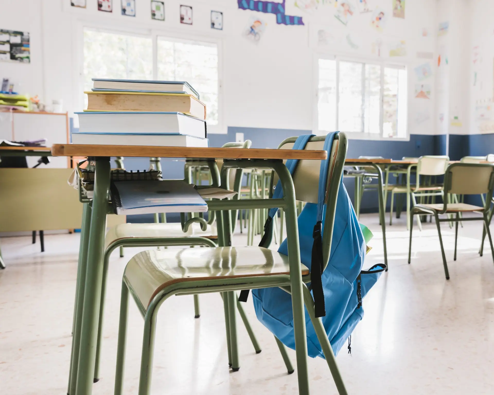 Ein leeres Klassenzimmer mit grünen Stühlen und Tischen, auf denen Bücher gestapelt sind; ein blauer Rucksack hängt an einem Stuhl. Schulreinigung bei Erdan Glas- und Gebäudereinigung.