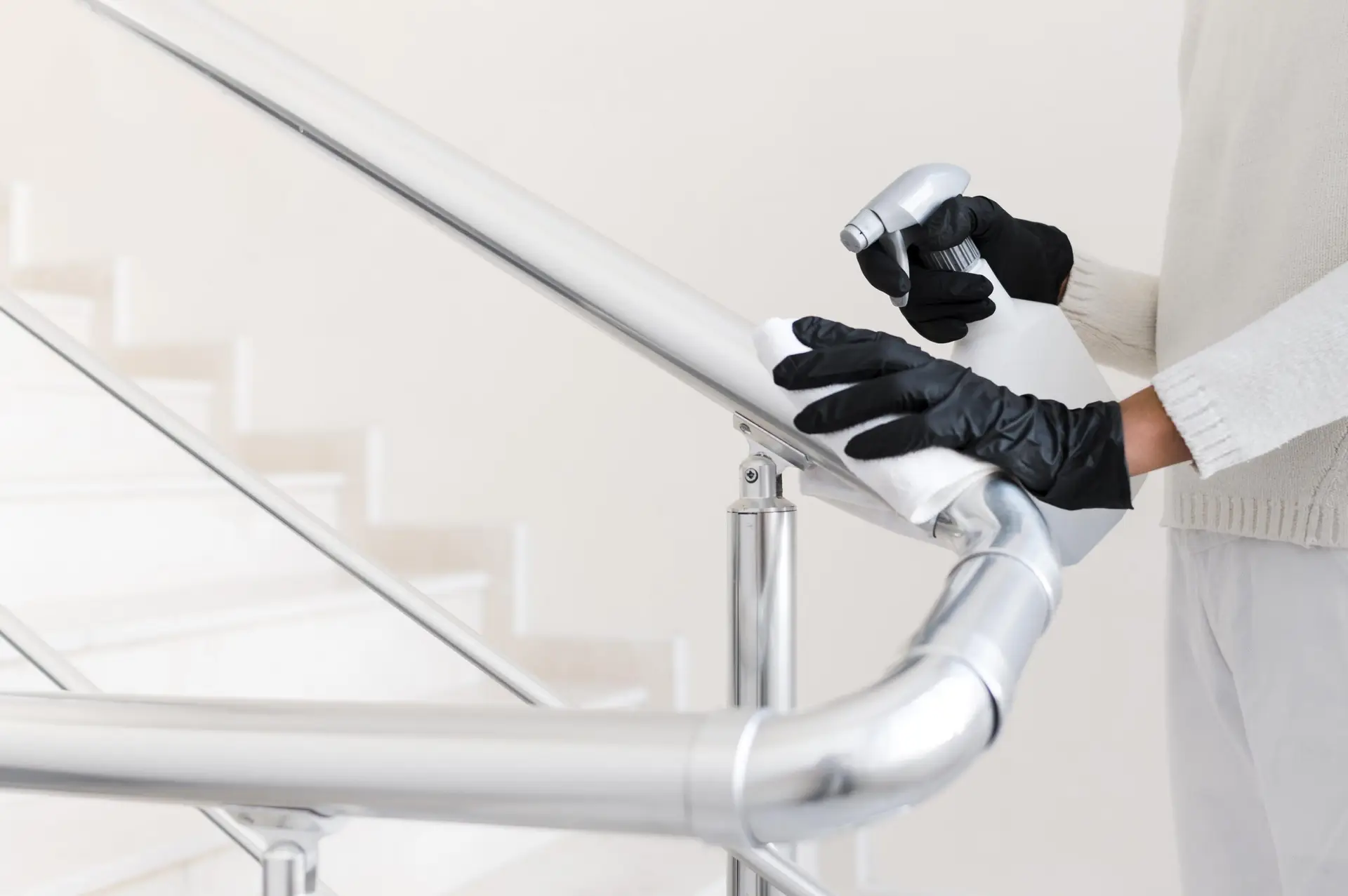 Eine Person mit schwarzen Handschuhen desinfiziert ein metallisches Treppengeländer mit einem Tuch und einem Spray. Treppenhausreinigung bei Erdan Glas- und Gebäudereinigung